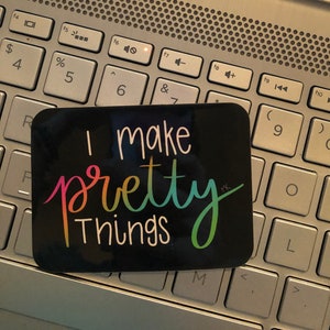 May include: A black sticker with rainbow lettering that reads "I make pretty things". The sticker is on a silver laptop keyboard.
