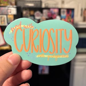 May include: A sticker with a light teal background and orange lettering that reads "lead with CURIOSITY not judgement". The sticker has a cloud-like shape and is held by a hand.