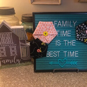May include: A teal blue letter board with the text "FAMILY TIME IS THE BEST TIME" in white letters. There is a heart with an arrow on the bottom of the board. There are three fabric hexagons with bee and floral patterns on the board.