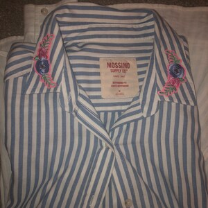 May include: A blue and white striped button-up shirt with embroidered floral details on the collar. The shirt has a label that reads "Mossimo Supply Co. Since 1987" and "Boyfriend Fit Corte Boyfriend".