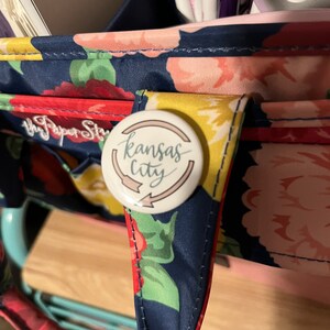 K&ouml;nnte beinhalten: Eine Tragetasche mit floralem Muster und einem Knopf mit der Aufschrift "Kansas City". Die Tasche hat einen marineblauen Hintergrund mit roten, rosa und gelben Blumen. Der Knopf ist wei&szlig; mit den Worten "Kansas City" in Schreibschrift.