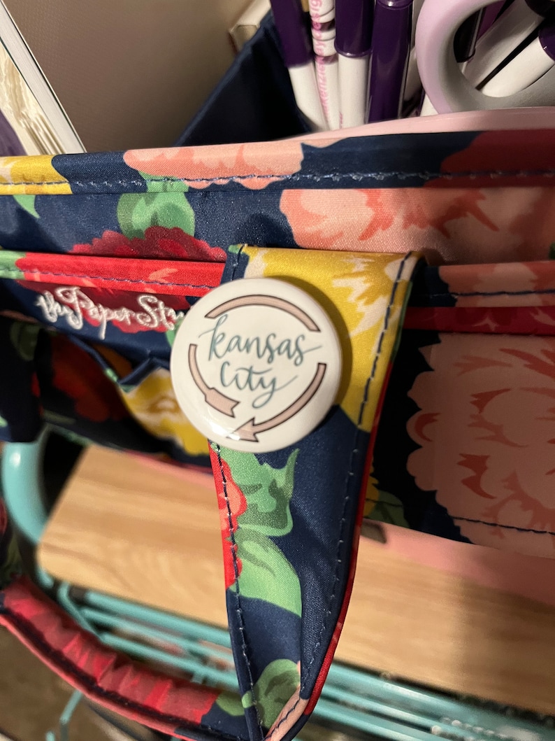 K&ouml;nnte beinhalten: Eine Tragetasche mit floralem Muster und einem Knopf mit der Aufschrift "Kansas City". Die Tasche hat einen marineblauen Hintergrund mit roten, rosa und gelben Blumen. Der Knopf ist wei&szlig; mit den Worten "Kansas City" in Schreibschrift.
