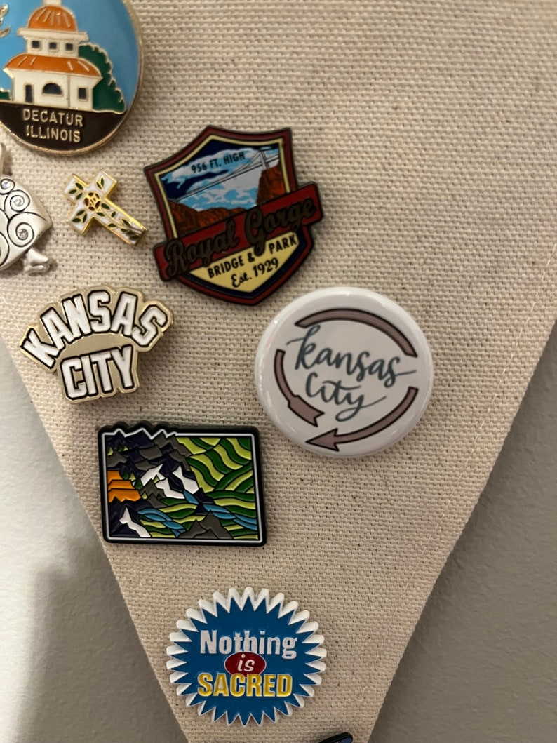 K&ouml;nnte beinhalten: Eine Sammlung von Emaille-Anstecknadeln und Buttons. Bemerkenswert sind eine Nadel mit "Kansas City" in Wei&szlig;, ein Button mit "Kansas City" in Schreibschrift und ein Badge mit dem Text "Nothing is Sacred". Weitere Nadeln zeigen Landschaften und Wahrzeichen.