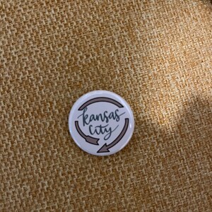 K&ouml;nnte beinhalten: Ein wei&szlig;er Ansteckbutton mit dem Schriftzug "Kansas City" in Schreibschrift, umgeben von zwei grauen Pfeilen. Der Button hat einen grauen Rand und liegt auf einem strukturierten, beigefarbenen Hintergrund.