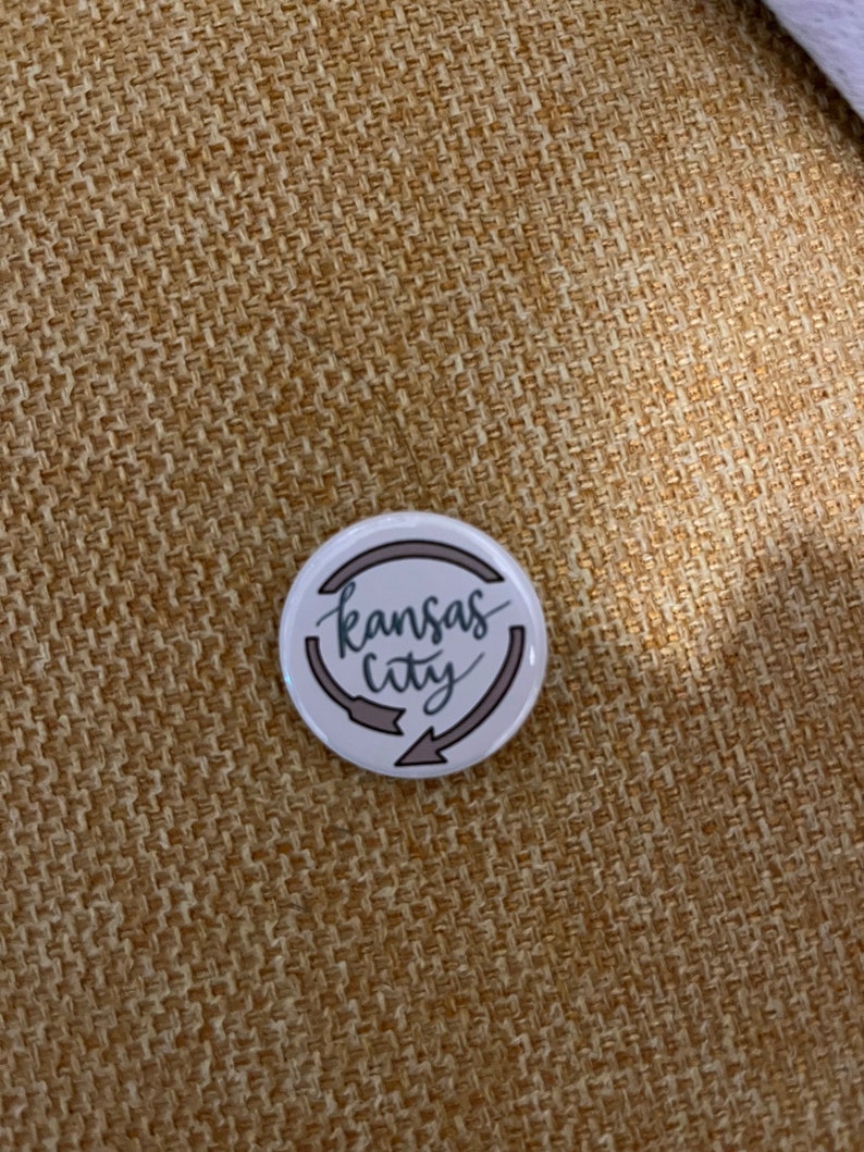 K&ouml;nnte beinhalten: Ein wei&szlig;er Ansteckbutton mit dem Schriftzug "Kansas City" in Schreibschrift, umgeben von zwei grauen Pfeilen. Der Button hat einen grauen Rand und liegt auf einem strukturierten, beigefarbenen Hintergrund.