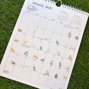 May include: A white wall calendar for January 2027 featuring a spiral binding at the top. The grid displays dates with small, colorful illustrations of various cats in different poses and activities scattered throughout the individual day squares.
