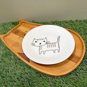 May include: A white ceramic plate with a cartoon cat illustration sits on a wooden cat-shaped tray. The plate features a brown striped cat design. The tray is made of light brown wood.