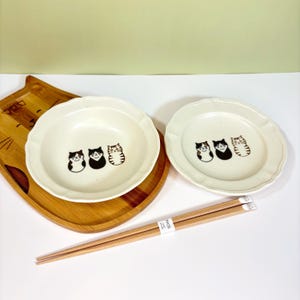 May include: Two off-white ceramic plates with scalloped edges, each adorned with three cartoon cat illustrations in black and brown. A wooden tray and chopsticks are also visible, creating a charming, cat-themed dining set.