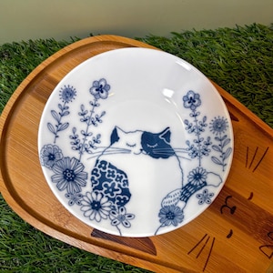 May include: White ceramic dish with a blue and white cat illustration surrounded by blue floral designs. The dish is round and sits on a wooden tray with a green background.