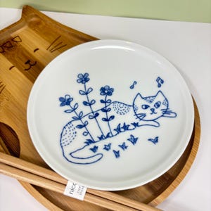 May include: White ceramic plate with a blue cat illustration, flowers, and musical notes. The cat is lying down among the flowers. The plate is on a bamboo tray with a cat face design and chopsticks.