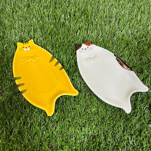 May include: Two ceramic cat-shaped plates. One plate is yellow with black stripes and the other is white with brown stripes.
