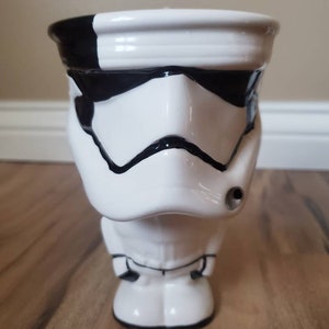 May include: White ceramic mug shaped like a Star Wars Stormtrooper. The mug has black details on the face and a black stripe around the top.