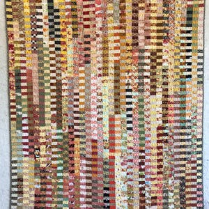 May include: A handmade quilt with a patchwork design. The quilt features vertical stripes of various fabrics in shades of yellow, brown, red, and green. The edges are finished with a black and white dotted border. A cozy, decorative textile.