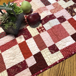 May include: A quilted table runner in shades of red, cream, and burgundy, featuring a patchwork design. The runner is adorned with various patterns and textures. A metal container with succulents and two pieces of fruit are on top of the runner.