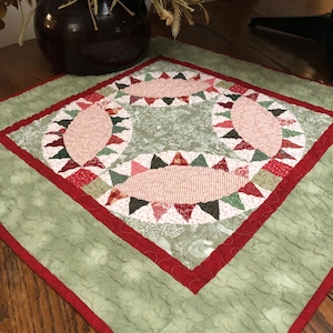 May include: A handmade, quilted table runner with a green background and a red border. The center features a design of pink ovals and red, green, and white triangles. The runner is placed on a wooden table, with a dark brown vase and dried flowers in the background.