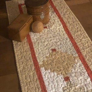 May include: A beige quilted table runner with a red border. The runner is decorated with a pattern of small white and brown squares. A wooden bucket with a lid and a wooden box are on top of the runner.