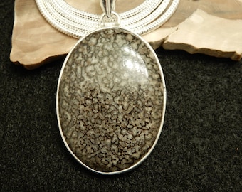 May include: Oval pendant with a silver-colored bezel. The pendant features a patterned, gray and brown stone. A silver chain is visible above the pendant, and the background is a dark gray.