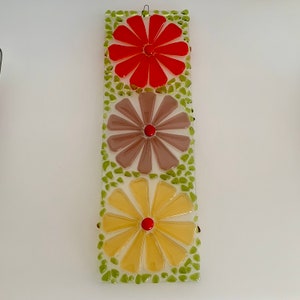 May include: A glass wall hanging with three mosaic flowers. The top flower is red, the middle flower is brown, and the bottom flower is yellow. Each flower has a red center.