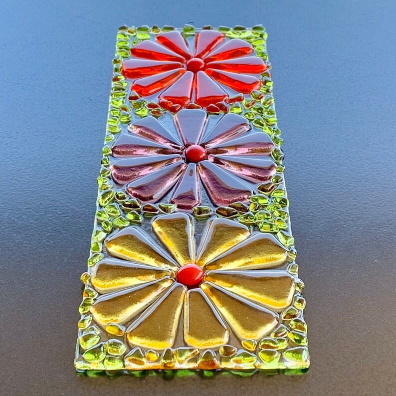 May include: A stained glass wall hanging with three colorful flowers. The flowers are made of glass pieces in shades of red, purple, and gold. The background is made of small green glass pieces.