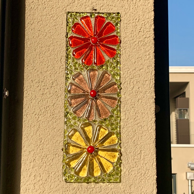 May include: A wall hanging with three glass flowers in a vertical row. The top flower is red, the middle flower is brown, and the bottom flower is yellow. Each flower has a red center.