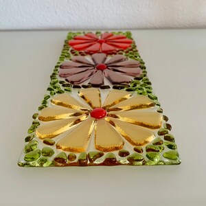 May include: A glass mosaic art piece featuring three stylized flowers. The flowers are made of small, colorful glass pieces and are arranged in a vertical row. The top flower is red, the middle flower is purple, and the bottom flower is gold. The flowers are surrounded by green glass pieces.