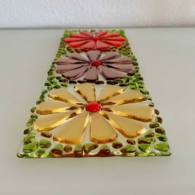 May include: A glass mosaic art piece featuring three stylized flowers. The flowers are made of small, colorful glass pieces and are arranged in a vertical row. The top flower is red, the middle flower is purple, and the bottom flower is gold. The flowers are surrounded by green glass pieces.