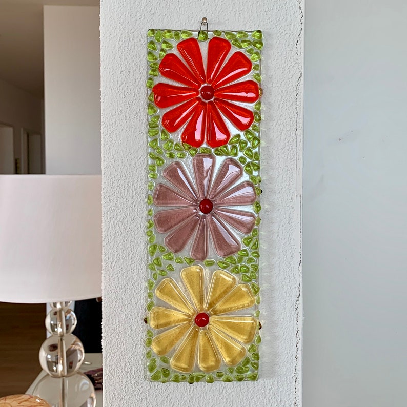 May include: A wall hanging with three glass flowers in shades of orange, purple, and yellow. The flowers are made of small pieces of glass and are set against a green background.