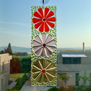 May include: A stained glass window hanging features three colorful flowers. The top flower is orange, the middle flower is pink, and the bottom flower is yellow. The flowers are surrounded by green glass pieces.