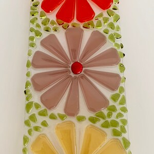 May include: A rectangular glass mosaic art piece with three stylized flowers. The top flower is red, the middle flower is light brown, and the bottom flower is yellow. The background is white with green glass pieces surrounding the flowers.