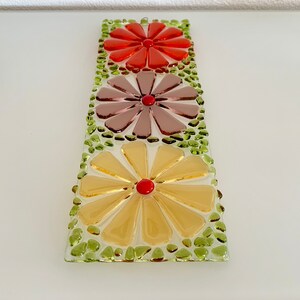 May include: A glass mosaic wall hanging with three flowers in shades of red, purple, and yellow. The flowers are surrounded by green glass pieces.