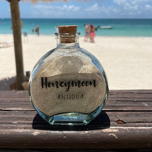 Pode incluir: Uma garrafa de vidro transparente cheia de areia, selada com uma rolha e rotulada "Honeymoon ANTIGUA". A garrafa está sobre uma superfície de madeira com uma praia e o oceano ao fundo sob um céu azul brilhante.