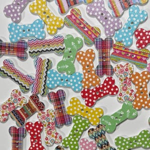 May include: A collection of bone-shaped buttons featuring various patterns including polka dots, plaid, chevron, and floral prints in multiple colors. Each button has two holes in the center for attachment, scattered across a plain white surface.