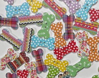 May include: A collection of bone-shaped buttons featuring various patterns including polka dots, plaid, chevron, and floral prints in multiple colors. Each button has two holes in the center for attachment, scattered across a plain white surface.