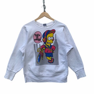 Könnte beinhalten: Weißes Sweatshirt mit einer Comicfigur mit Kopfhörern, Fahrrad und dem Text "Take it easy!". Die Figur trägt eine rote Jacke, blaue Shorts und eine "Brooklyn"-Kappe. Das Design beinhaltet eine Stadtkulisse.