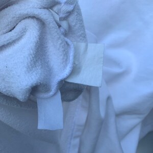 May include: Close-up of a white terry cloth garment, possibly a robe or towel, with a tag attached. The fabric is soft and textured, with visible loops. The garment is folded, revealing the inside and the attached tag.