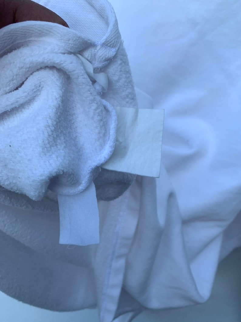 May include: Close-up of a white terry cloth garment, possibly a robe or towel, with a tag attached. The fabric is soft and textured, with visible loops. The garment is folded, revealing the inside and the attached tag.