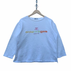 May include: White long-sleeve sweatshirt with the text "courr&egrave;ges paris" embroidered in orange, yellow, and red. The sweatshirt has a crew neck and a small split at the bottom of each side. The garment is hanging from a wooden hanger.