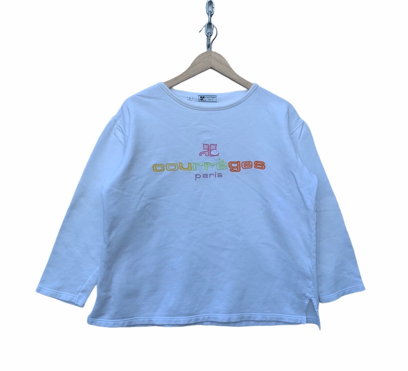 May include: White long-sleeve sweatshirt with the text "courr&egrave;ges paris" embroidered in orange, yellow, and red. The sweatshirt has a crew neck and a small split at the bottom of each side. The garment is hanging from a wooden hanger.