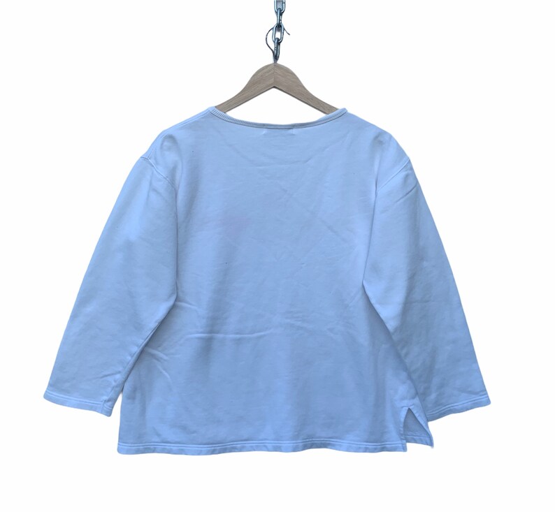 May include: A white long-sleeve shirt with a scoop neck and side slits. The shirt is hanging on a wooden hanger suspended by a metal chain. The shirt is made of a soft, lightweight material.