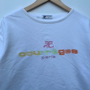 May include: White crew neck sweatshirt with the embroidered text "courr&egrave;ges paris" in yellow, green, and orange. The sweatshirt has a small logo on the upper left and is hanging on a wooden hanger.