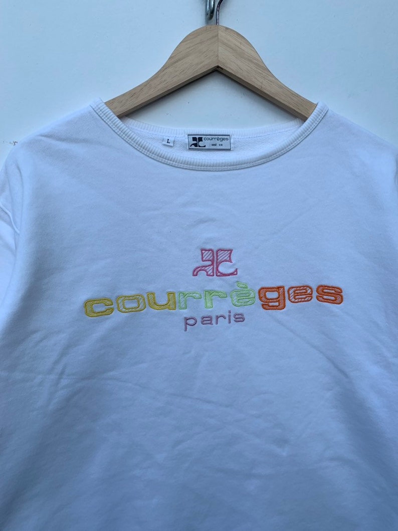 May include: White crew neck sweatshirt with the embroidered text "courr&egrave;ges paris" in yellow, green, and orange. The sweatshirt has a small logo on the upper left and is hanging on a wooden hanger.