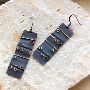 May include: A pair of black and copper earrings with a rectangular shape and a textured surface. The earrings have multiple small holes and a wire hook.