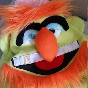 May include: A whimsical green hat with an orange nose, orange shaggy hair, and a wide, toothy grin. The hat features large black eyebrows, blue eyes, and a red mouth. A pink fluffy detail is visible on the top of the hat.