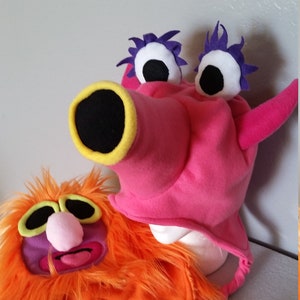 May include: Two plush character hats. One is orange with a furry texture, purple accents, and a braided tail. The other is pink with large white and black eyes, purple accents, and a large black and yellow snout.