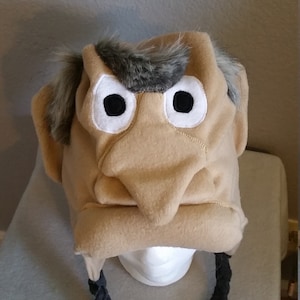 May include: A novelty hat shaped like a cartoon face, featuring a large nose, black eyes, and gray hair. The hat is tan with black braids hanging down. The hat is displayed on a white mannequin head.