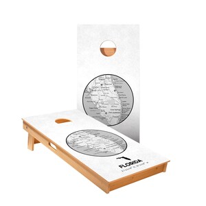 May include: A white cornhole board with a black and white map of Florida on it. The board is made of wood and has a brown stain. The map is centered on the board and includes the state's major cities and highways. The text "FLORIDA" and the coordinates "27.6648° N, 81.5158° W" are printed below the map.