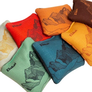 May include: Six bean bags with different colored fabric and a mountain range design. The bean bags are labeled "Elakai" and are arranged in a semi-circle.