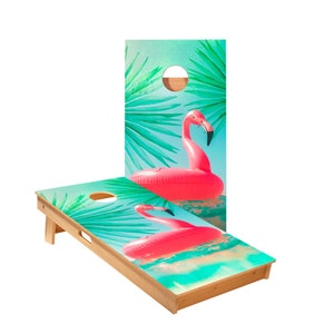 May include: A wooden cornhole game with a tropical design featuring a pink flamingo pool float on a turquoise background with palm leaves.