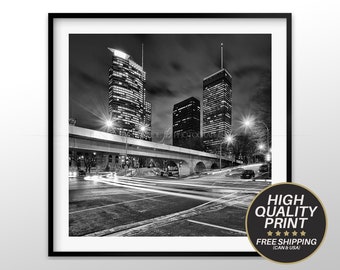 Puede incluir: Fotografía en blanco y negro de un horizonte urbano de noche. La imagen muestra tres edificios altos con ventanas iluminadas, un puente y una calle con coches que pasan. Las farolas están encendidas y los faros de los coches crean rayas de luz en la foto.