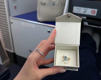 May include: A white ring box with a blue gemstone ring inside. The ring is set in gold and has a teardrop shape. The box is open and the ring is visible.